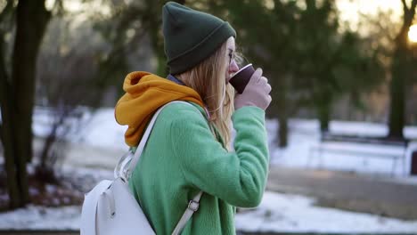 Woman-walking-with-hot-drink-by-winter-city-park-or-forest,-enjoy-weather,-side-view