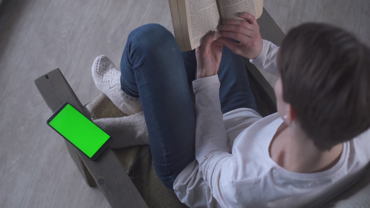 niña con cabello corto en vaqueros y calcetines de lana se sienta en casa en un sillón y lee un libro en una mañana de invierno. cerca de un teléfono móvil pantalla verde. clave de croma teléfono celular pantalla verde nuevo concepto de tecnología.