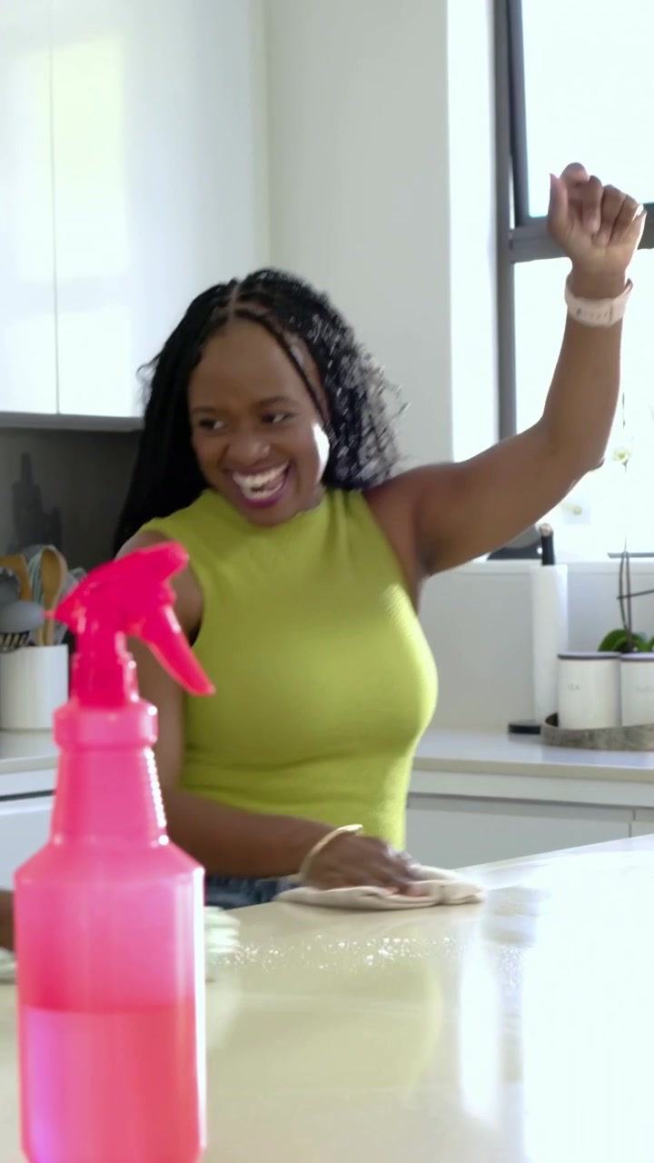 Video verticale: Una donna con top color lime spruzza e pulisce la farina rovesciata sull'isola della cucina con una bottiglia blu.