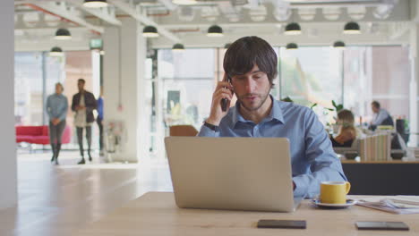 Businessman-Sitting-At-Desk-On-Phone-Call-In-Modern-Open-Plan-Office-With-Colleagues-In-Background