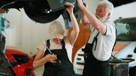 una mujer adulta y un viejo mecánico trabajan en la renovación de un coche dañado en un taller quitando un parachoques viejo