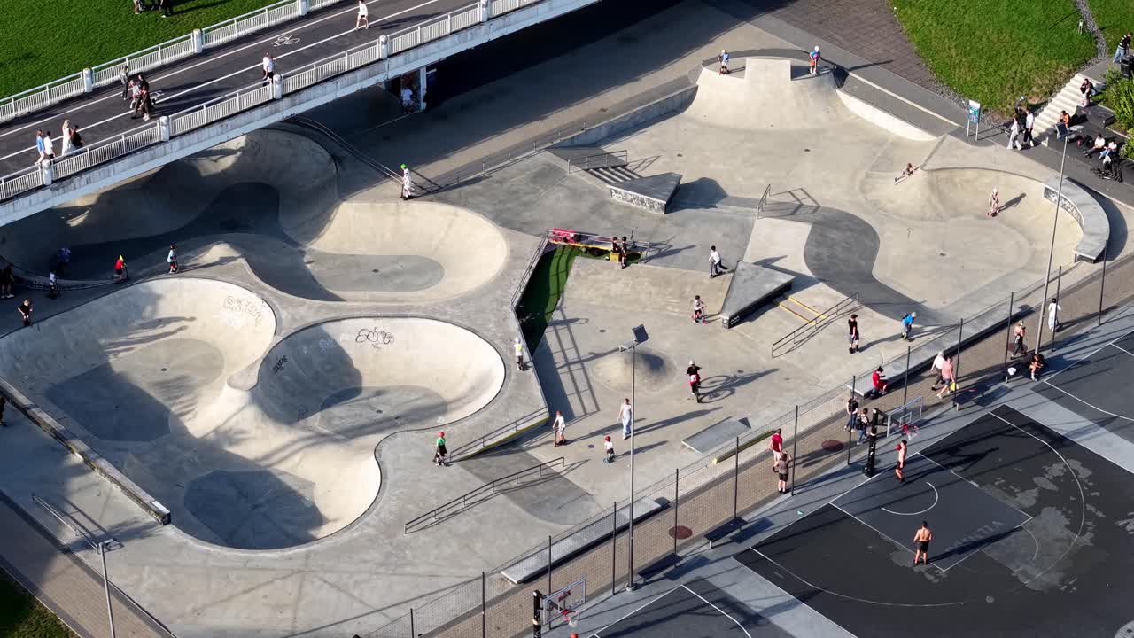 Aerial view of Vilnius skatepark by river bridge with skateboard, BMX and nearby basketball courts on sunny day, perfect for youth culture and urban leisure