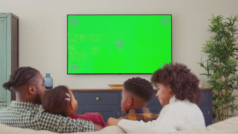 Vista-Trasera-De-Una-Familia-Sentada-En-Un-Sofá-En-Casa-Viendo-Televisión-De-Pantalla-Verde-Y-Riéndose-De-La-Comedia---Filmada-En-Cámara-Lenta