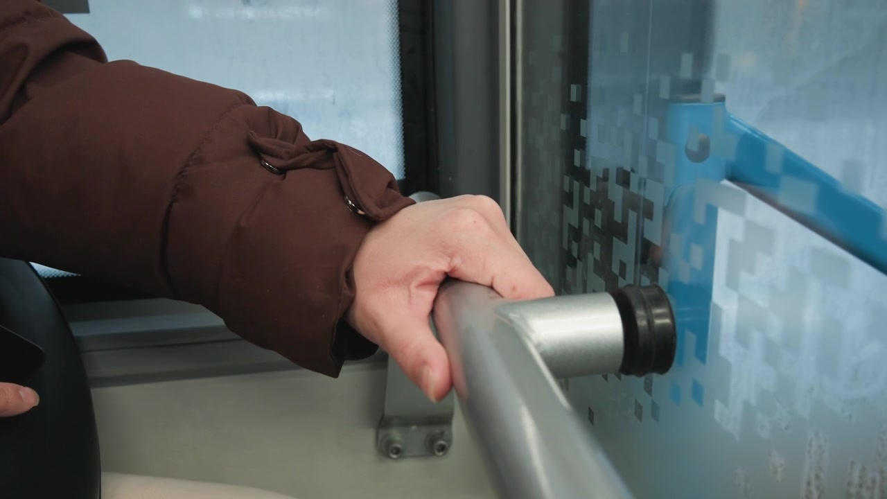 Human hand grips cylindrical handle on frosted glass partition inside public transit interior, with visible pulling motion, gentle release, diffuse daylight, and soft background seating
