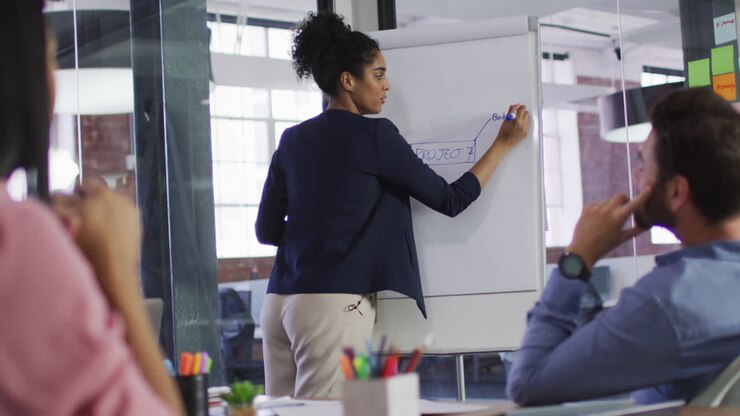 Femme métisse debout devant un tableau blanc faisant une présentation à un groupe diversifié de collègues