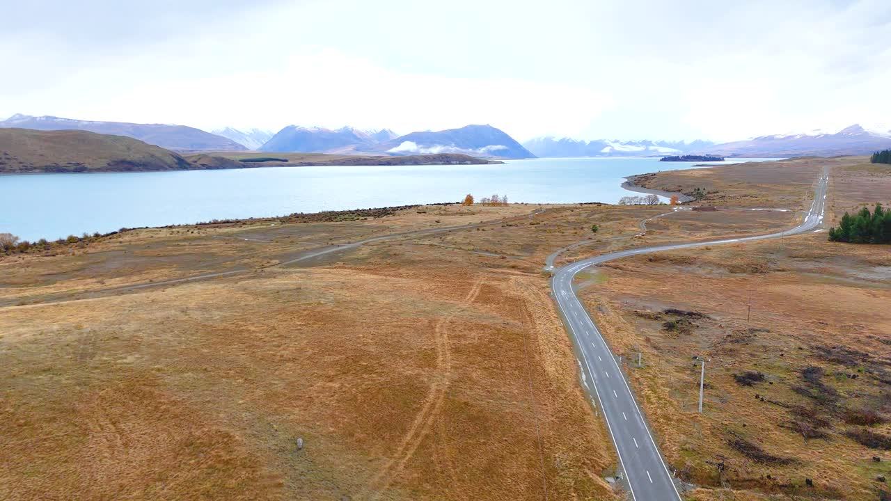 Aerial footage captures a serene road beside Lake Tekapo, showcasing expansive landscapes under soft, natural lighting