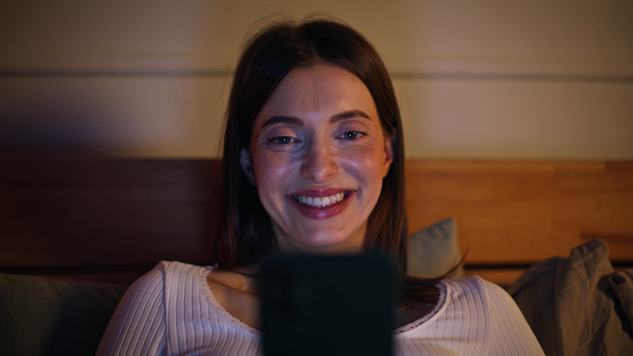 Nærbillede af en glad pige, der læser SMS på sin mobiltelefon, mens hun ligger i en hyggelig seng. Smilende kvinde.