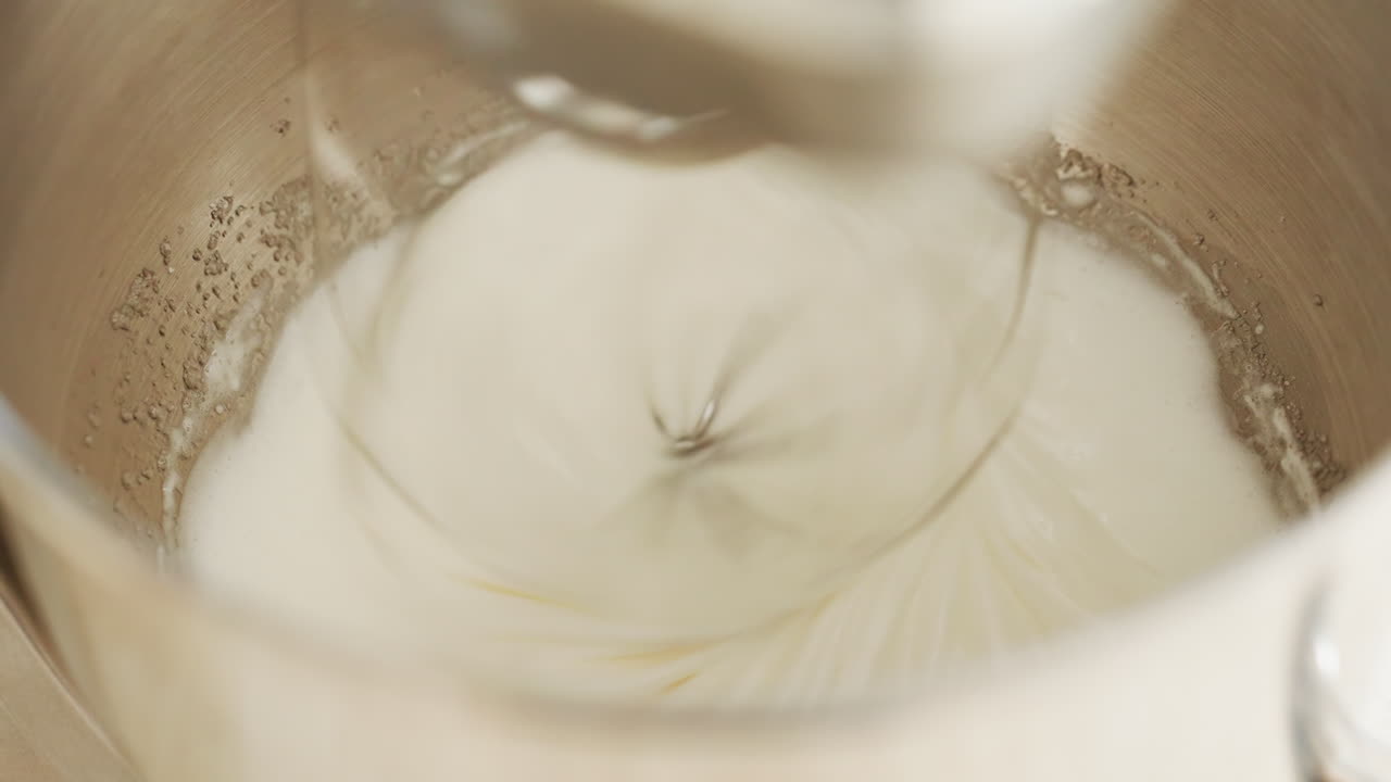 Close up of electric stand mixer blending flour and liquid in stainless steel bowl to form dough, showing smooth mixing motion and swirling ingredients for baking