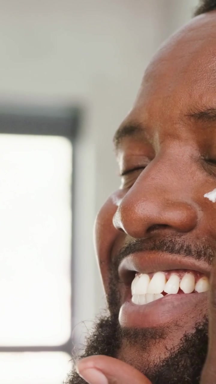 Video verticale: Uomo barbuto che si massaggia, applicando crema su un punto della guancia vicino alla finestra per la cura, con la mano destra.