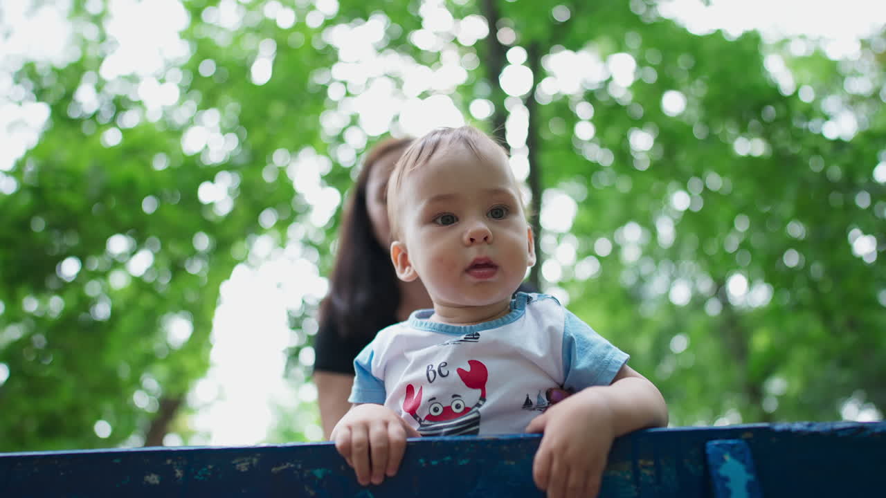 Curious lovely child outdoors. Baby boy standing on the bench looking at camera. Mother's behind her son. Low angle view.