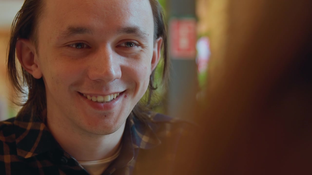 Close up of young man with long hair wearing plaid shirt speaking and smiling in indoor setting. scene shows soft lighting and warm tones focusing on subject facial expressions