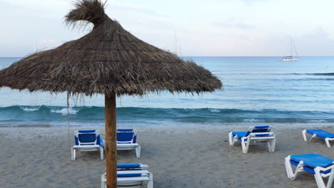 Rearview-of-an-empty-beach-at-dawn-with-sunbeds-and-straw-umbrellas,-small-waves-on-the-sea