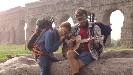 happy young couple backpackers tourists sitting on a log trunk playing guitar singing in front of ancient roman aqueduct ruins in romantic parco degli acquedotti park in rome at sunrise sleeping bag slow motion