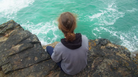depressed young person sitting on coastal cliff edge watching the ocean seeking peace and calmness