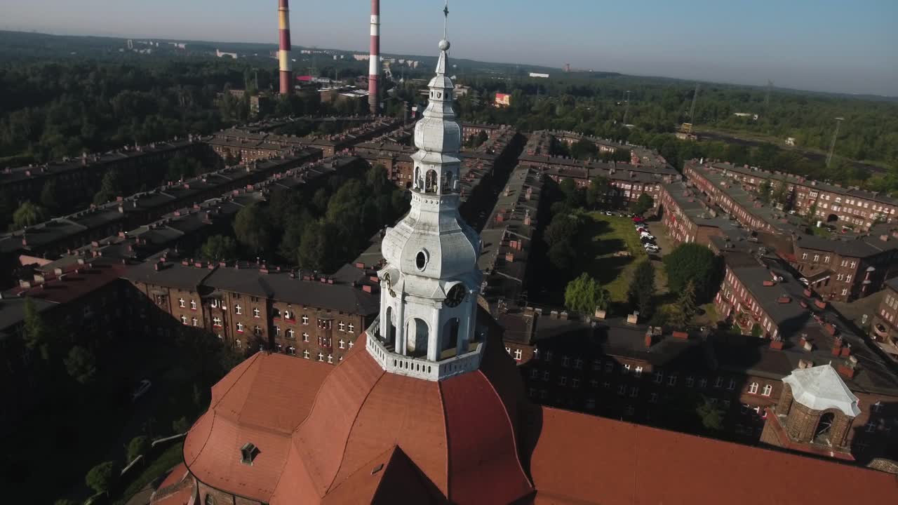 Tower of a historic church located in Nikiszowiec, a preserved industrial settlement. Factory chimneys rise in the distance against a clear blue summer sky, 4K drone shot