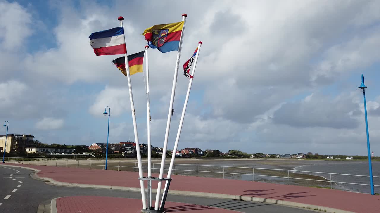 Flagstaffs with flags of Germany, Schleswig-Holstein, Amrum are waving in heavy seaside wind, clounds rolling by in the background, The whole flagstaff is visible, from top to bottom