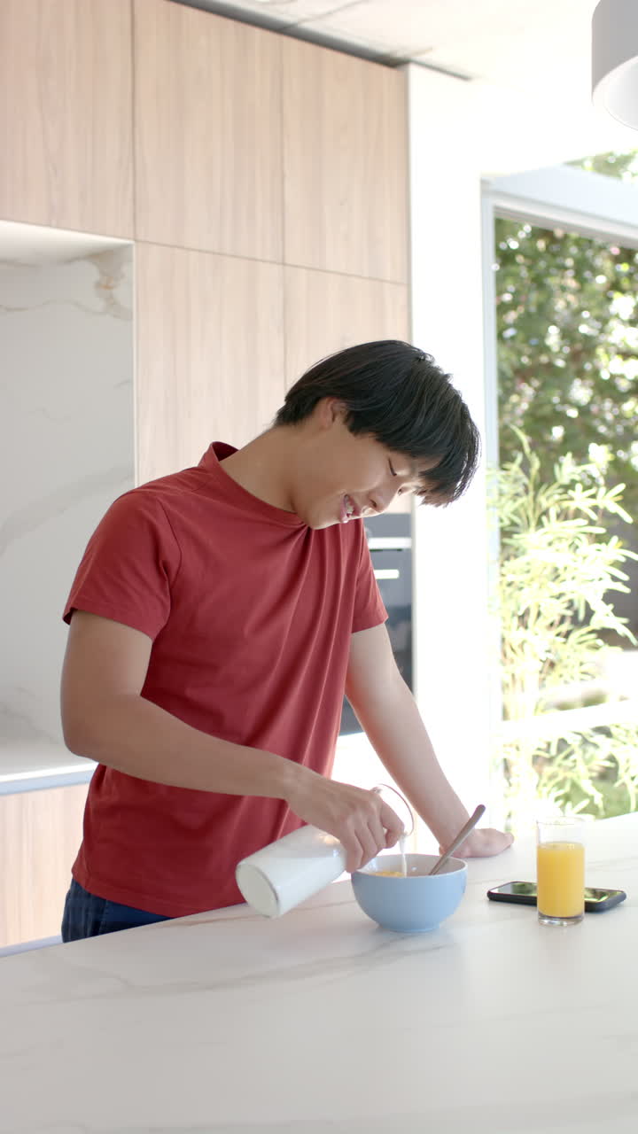 Vertical video: Asian teenage boy pouring milk into cereal bowl in kitchen, preparing breakfast at h