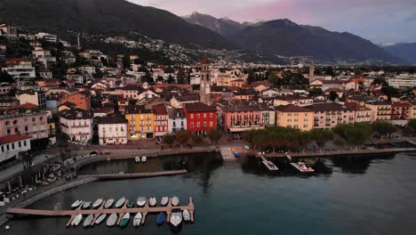 Sobrevuelo-Aéreo-Sobre-Las-Orillas-Del-Lago-Maggiore-Hacia-El-Paseo-Marítimo-Y-La-Torre-De-La-Iglesia-De-Ascona,-Suiza,-Con-Vistas-A-Las-Montañas,-Barcos,-Casas-Coloridas