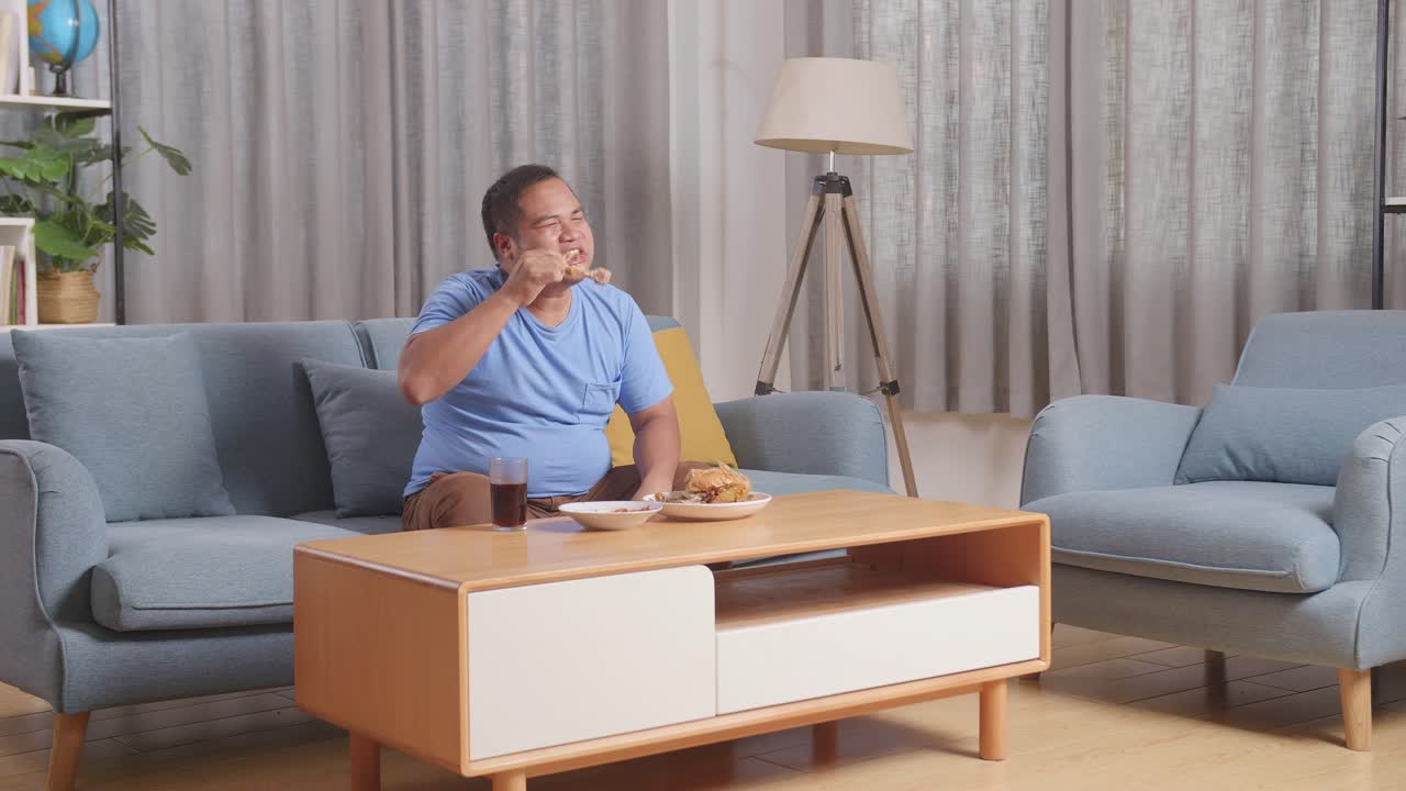 한 아시아 남자의 몸이 가득 차 있고, 패스트푸드를 먹고, 고기를 먹고, 배를 만지기 전에, 집에서 거실에 있는 소파에 앉아 있다.