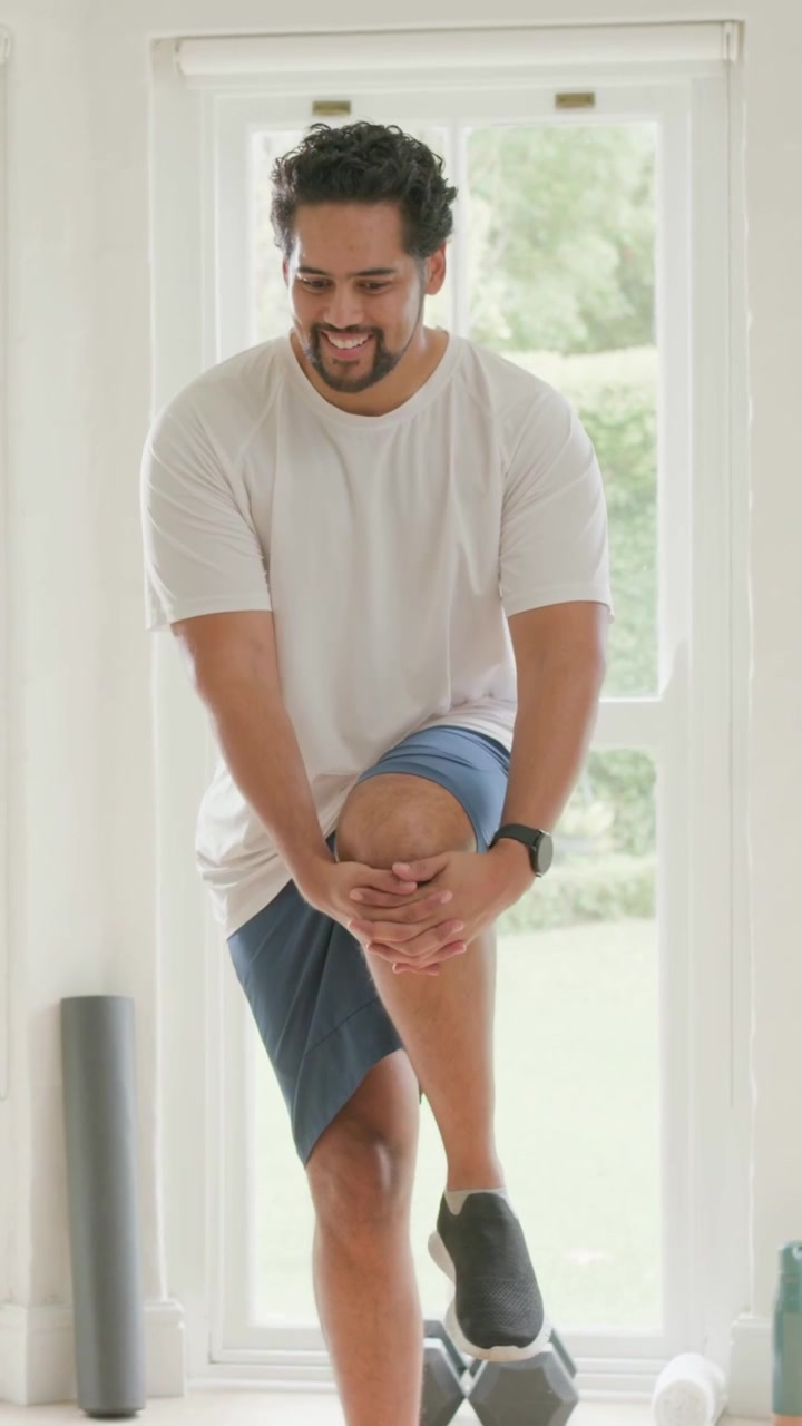 Vertical video: Warming up, man doing knee lifts at home by door, wearing tee, watch