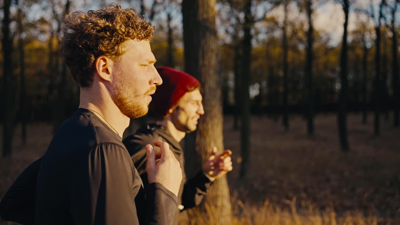 A brunette man with curly hair and a beard in black sportswear runs with his partner through the morning sunny autumn forest during his jog