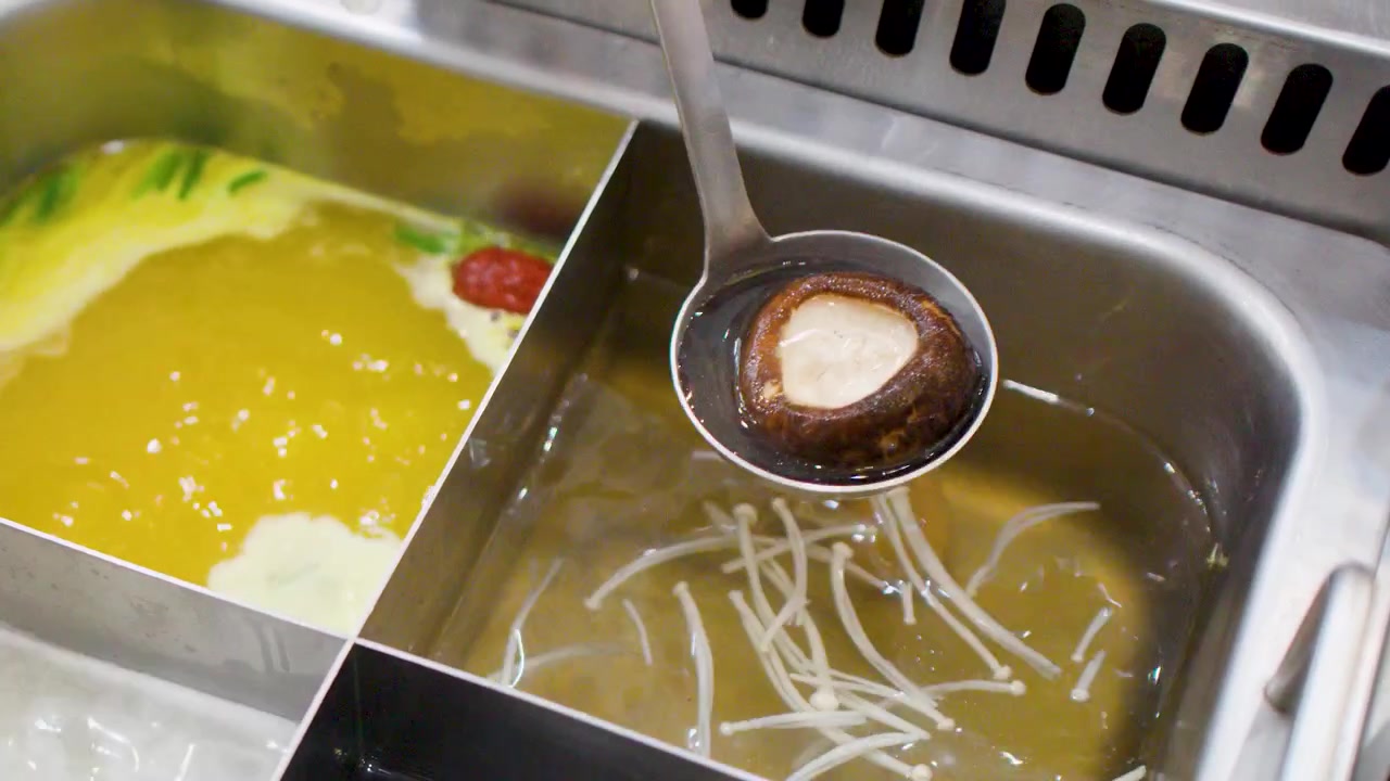 Un mestolo di metallo solleva un fungo shiitake da un brodo bollente trasparente in una pentola divisa