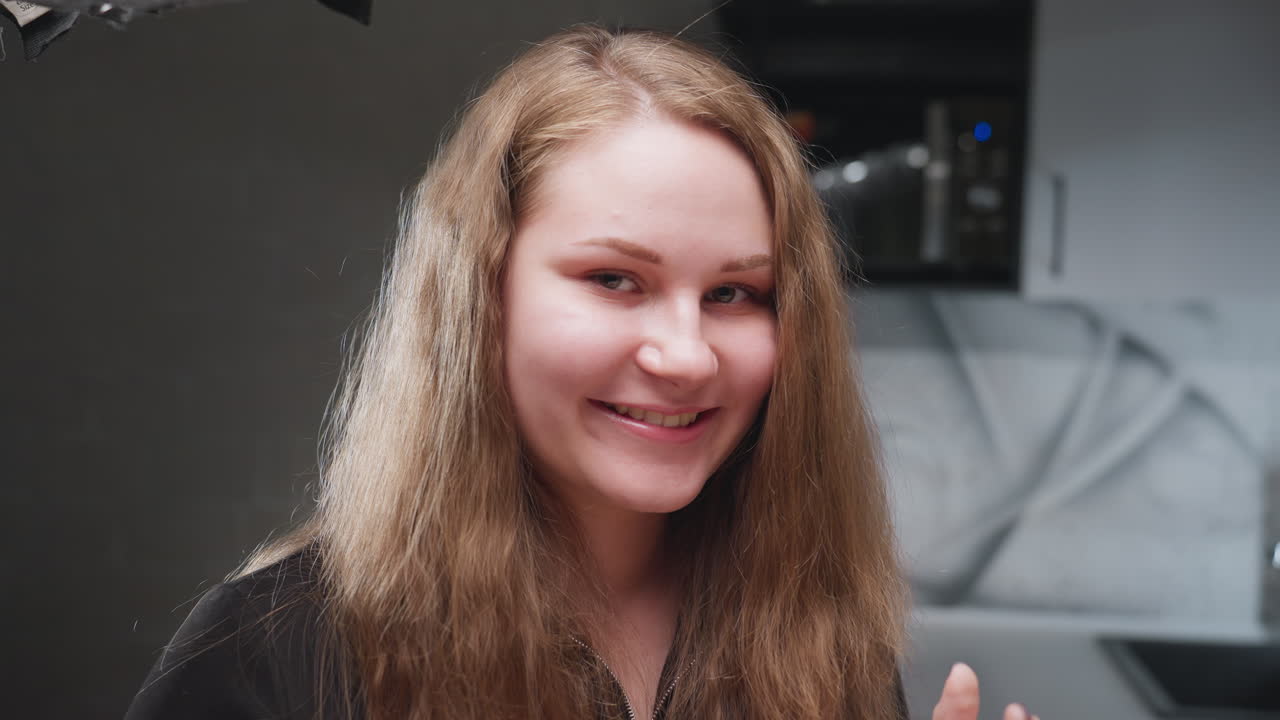 Portrait view young woman with long natural hair standing in modern kitchen, gentle smile, relaxed posture, soft light on face, clean cabinets, blurred background, cozy home mood, lifestyle scene