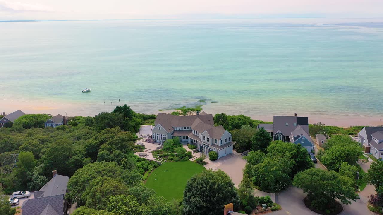 A Cape Cod home faces sandy beach where families stroll the coast, turquoise water glimmers in gentle late-day sunlight, and trees add shade to the shore