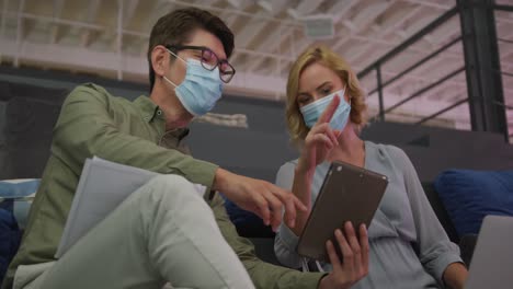 Asian-man-and-caucasian-woman-wearing-face-masks-discussing-over-a-digital-tablet-at-office