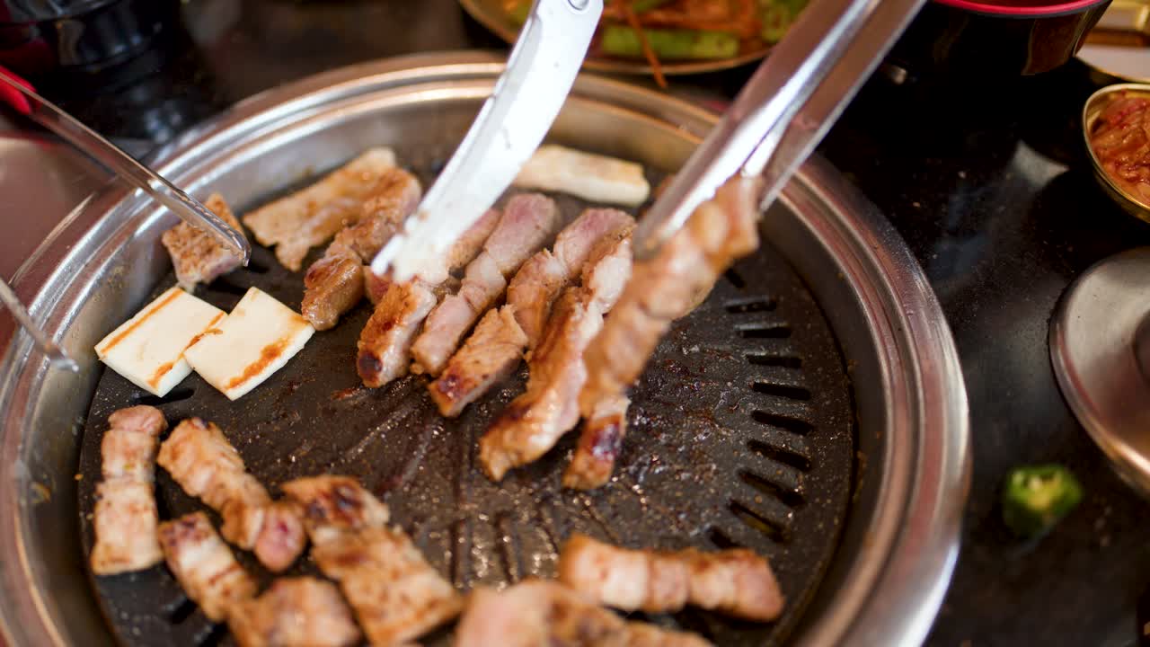 Hand uses tongs to flip pork belly slices on tabletop Korean barbecue grill, overhead view