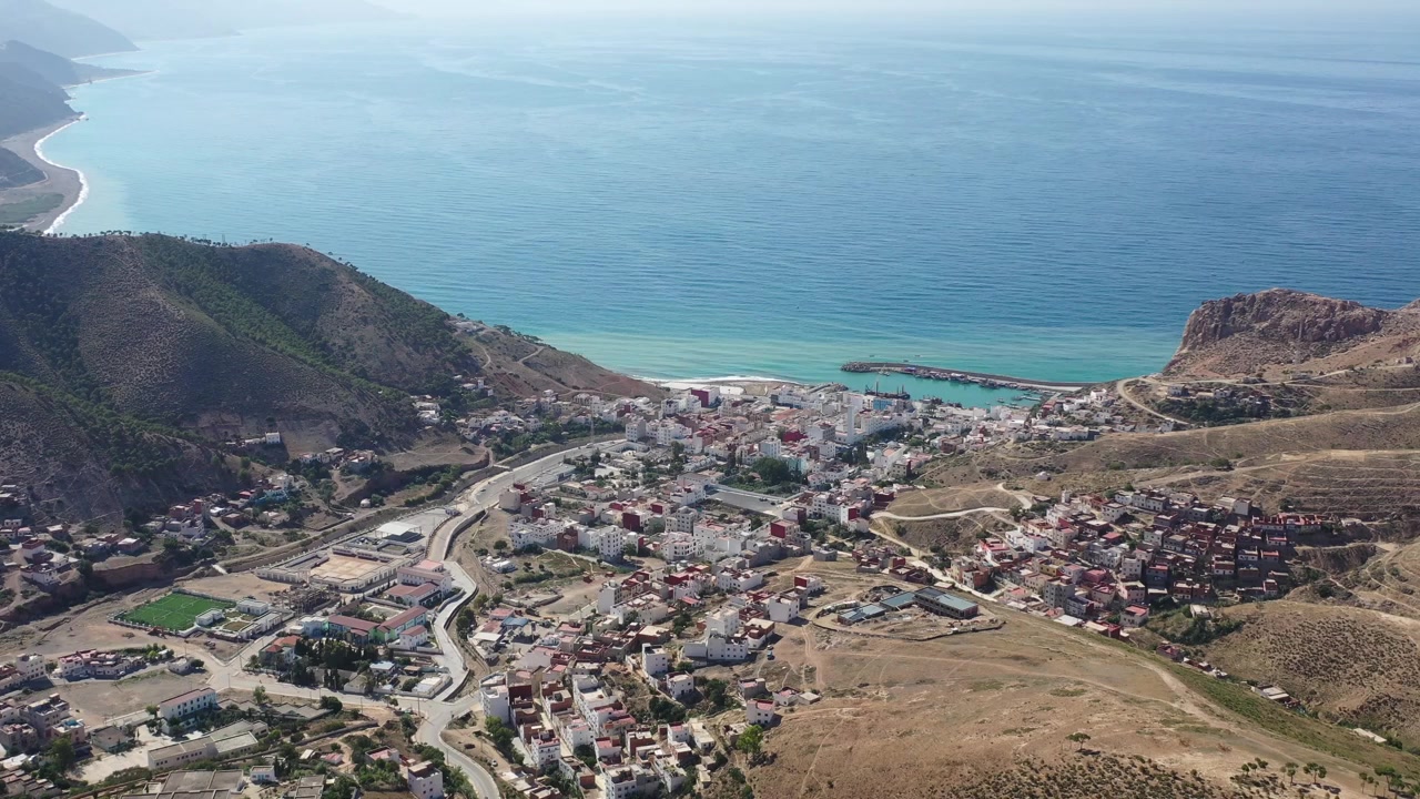 Ampia vista aerea di una città costiera che si estende attraverso una valle montuosa aspra verso il Mar Mediterraneo nel Marocco settentrionale, con strade tortuose, colline e un piccolo porto