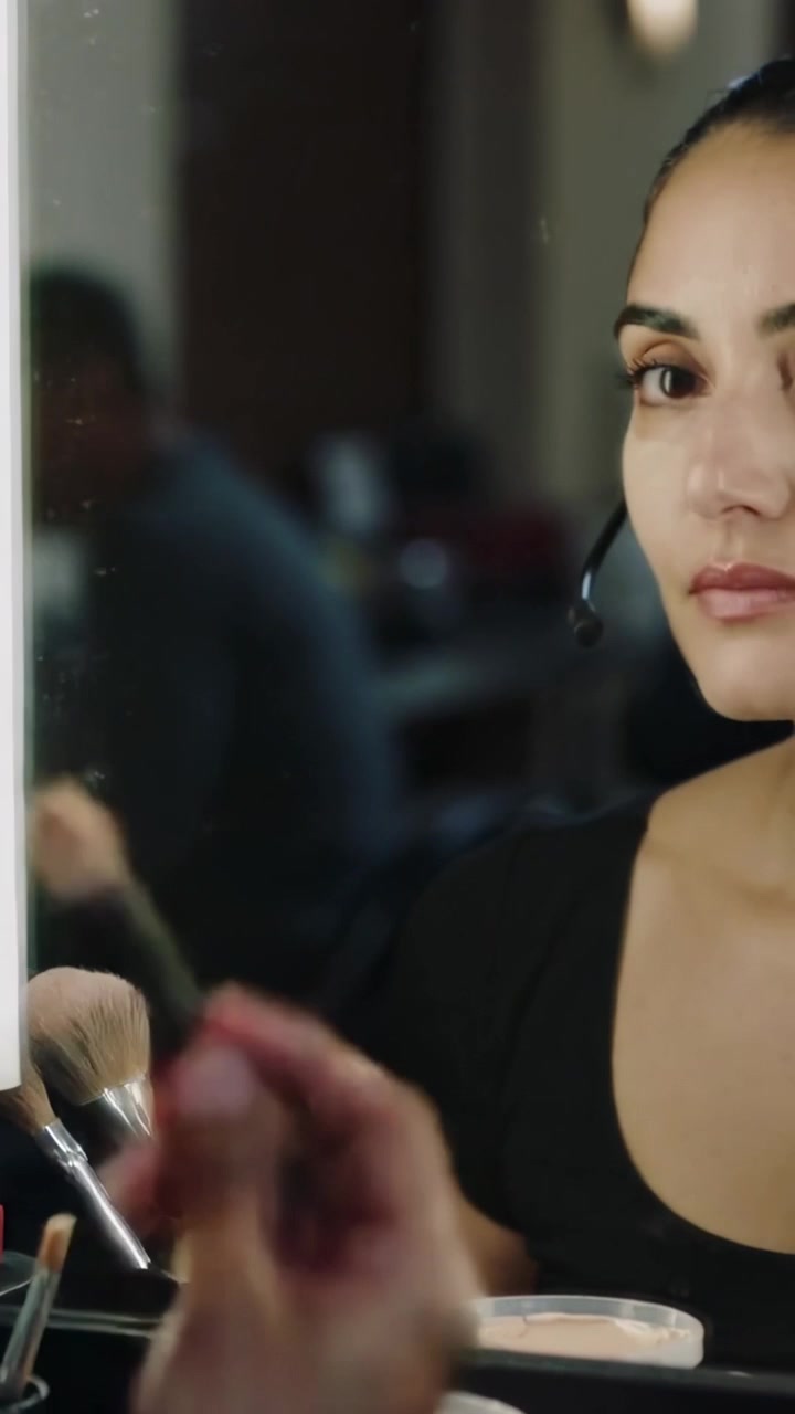 Video verticale: Donna in nero con cuffie che si trucca davanti a uno specchio illuminato per una chiamata, con spazio per il testo.