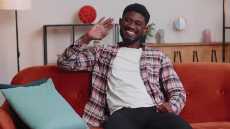 young man smiling friendly at camera, waving hands gesturing hello, hi, greeting at home on sofa