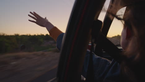 Frau-Im-Auto-Hält-Die-Hand-Aus-Dem-Fenster-Und-Spürt,-Wie-Der-Wind-Durch-Die-Finger-Weht,-Während-Sie-Auf-Einem-Roadtrip-Durch-Die-Landschaft-Fährt-Und-In-Den-Sommerurlaub-Reist-Und-Bei-Sonnenaufgang-Die-Freiheit-Auf-Der-Straße-Genießt