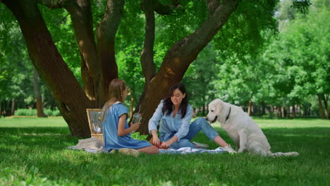 Fröhliche-Mutter-Füttert-Labrador-Beim-Picknick.-Mutter-Sitzt-Mit-Tochter-Auf-Einer-Decke