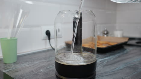 close-up of water being poured into a glass jar with a filter, the blurred background features a kitchen wall socket, tray with cookies, and a cup