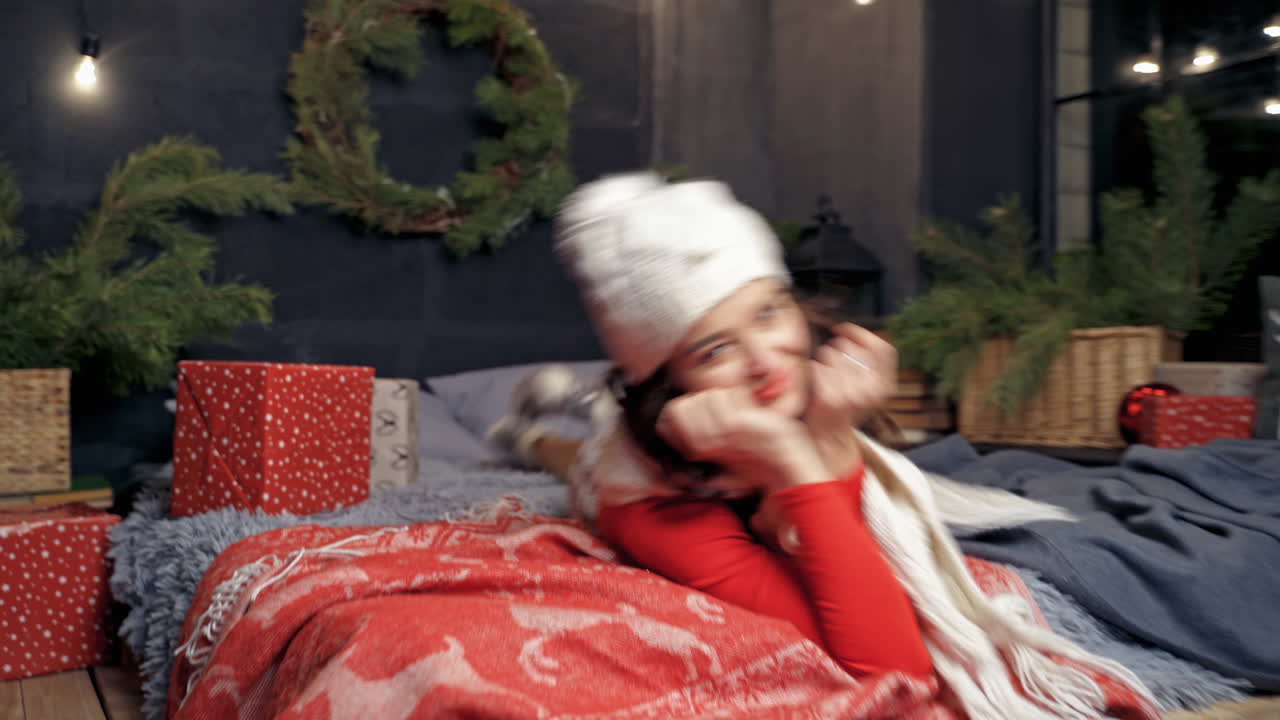 Emotional girl at Christmas. Beautiful young woman in knitted cap and scarf laying on bed on Christmas presents and fir branches background.
