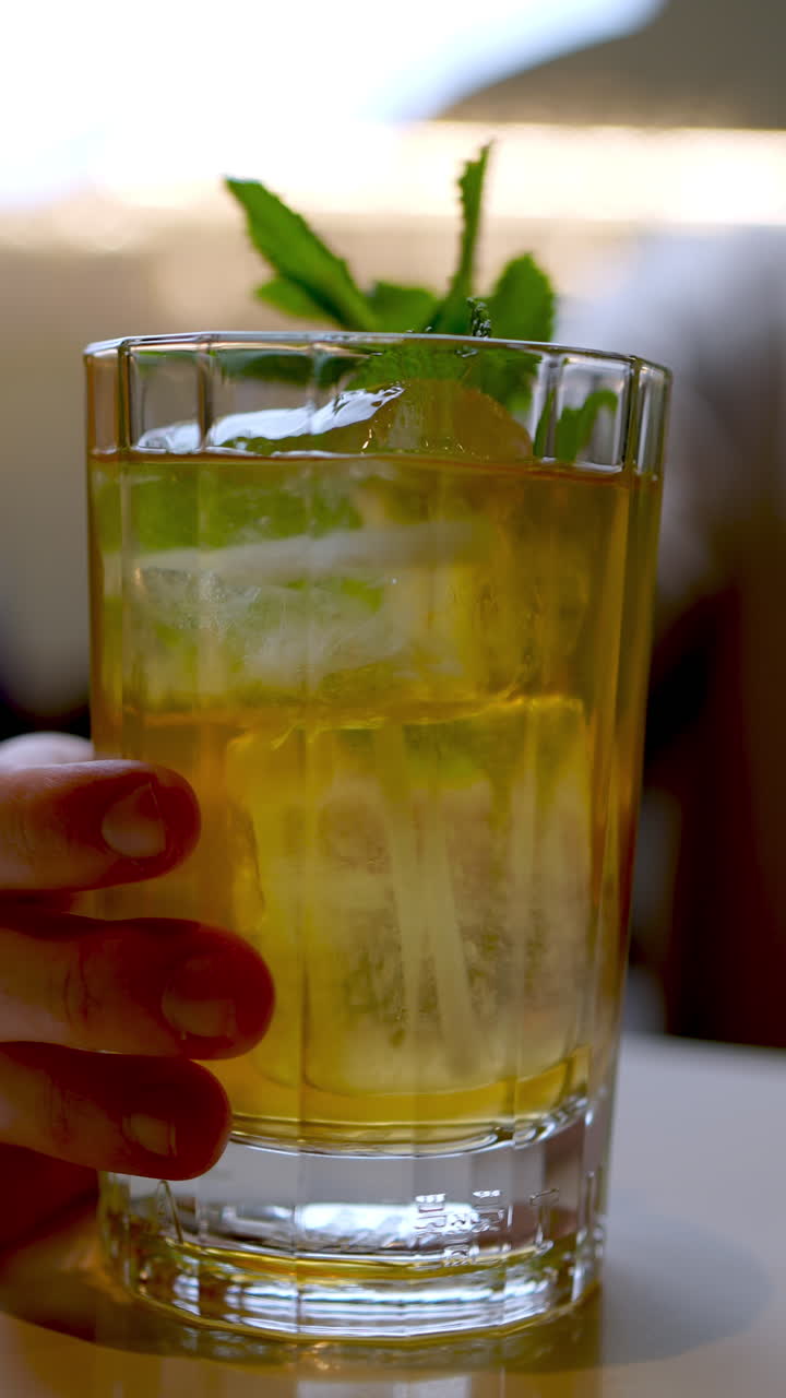 Vertical footage Hand delicately grasping transparent glass, showcasing refreshing iced tea with green mint leaves and sparkling ice cubes against pristine white background