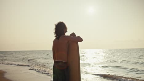 Rear-view-of-a-guy-with-medium-length-hair-with-a-bare-torso-in-shorts-stands-and-holds-his-wooden-yellow-surfboard-on-a-sandy-rocky-shore-near-the-sea-at-Sunrise-in-the-morning