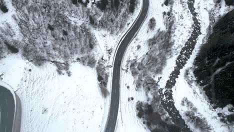 Aerial-top-down-view-of-a-small-car-driving-on-a-snow-covered-mountain-road-during-a-cloudy-winter-day-with-snowfall-in-Switzerland