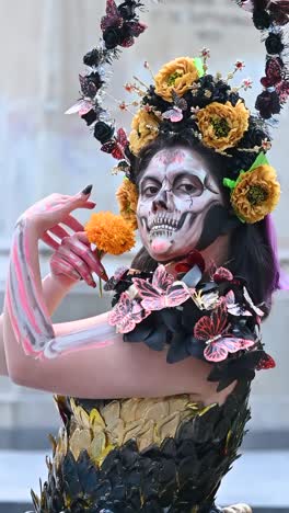 a woman with detailed makeup depicting a catrina, adorned with a large flower crown and holding a marigold, celebrates the day of the dead in mexico