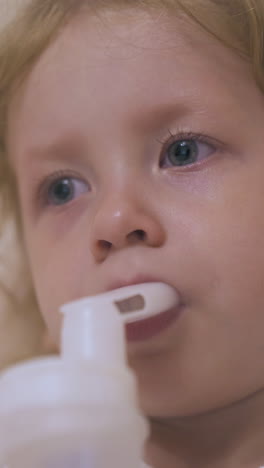 pretty little girl has procedure with modern inhaler lying in soft bed with pillow at home extreme close view