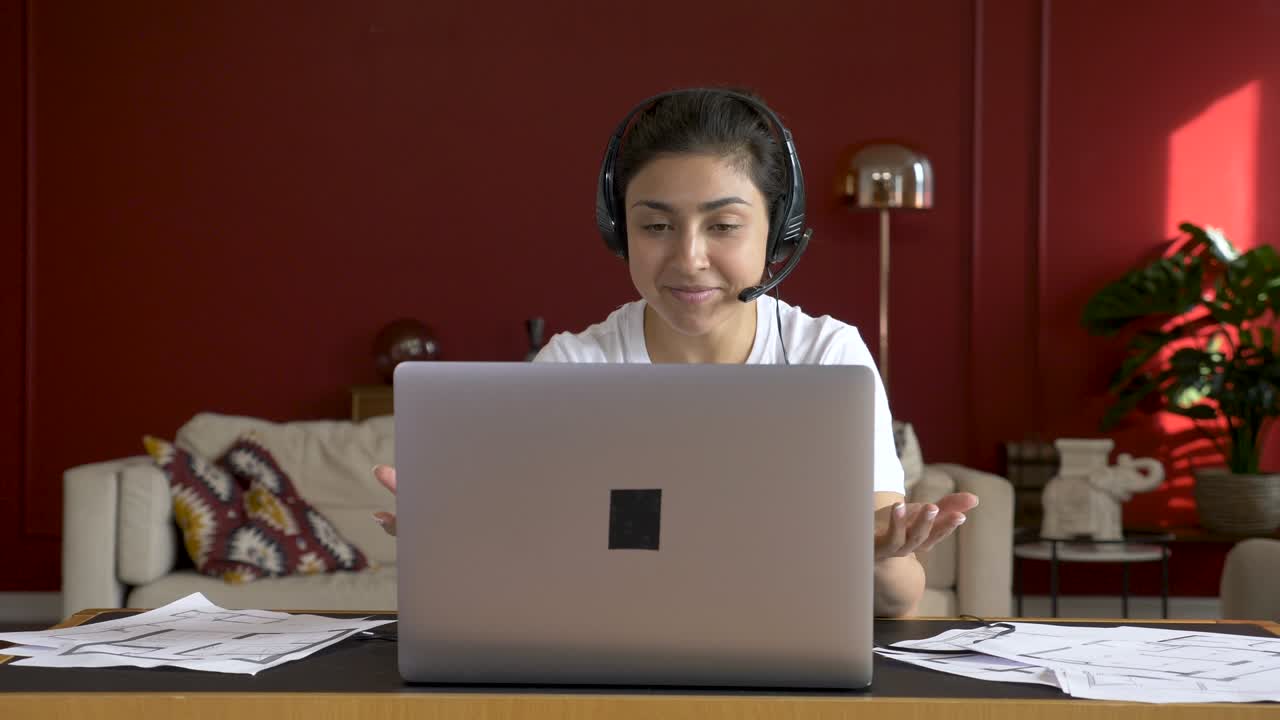 Young indian woman wears headset conference calling on laptop talks with online teacher studying, working from home. Lady student e learning using computer webcam chat makes notes. Distance education concept