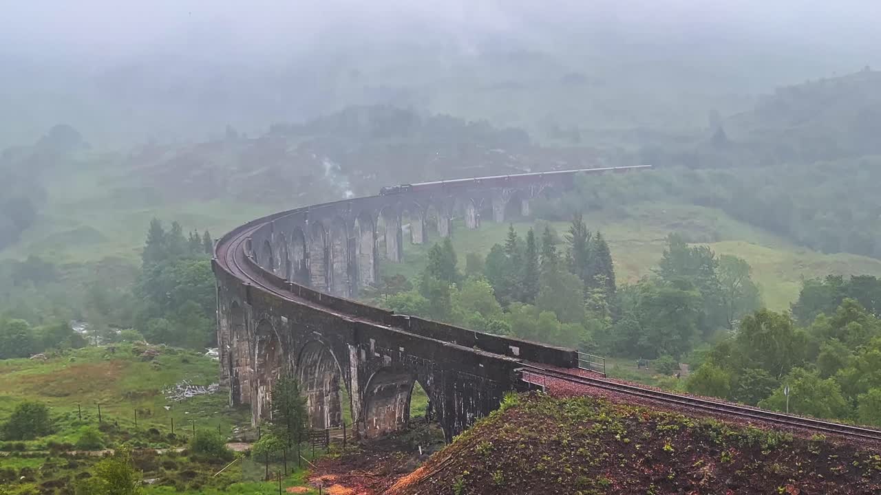 Steam Train Traveling Through Old And Historic Glenfinnan Viaduct On A Misty And Rainy Day In Scotland. - wide shot