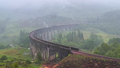 steam train traveling through old and historic glenfinnan viaduct on a misty and rainy day in scotland. - wide shot