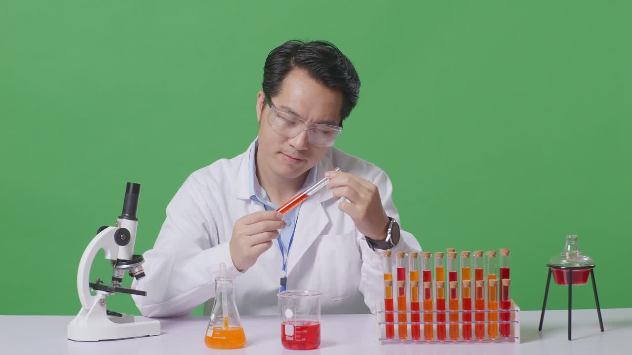 Asian Man Scientist Looking At The Red Liquid In The Test Tube Carefully While Working On The Table With Microscope In The Green Screen Background Laboratory