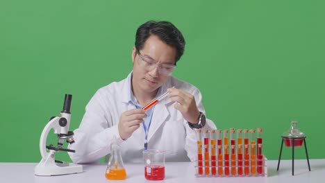asian man scientist looking at the red liquid in the test tube carefully while working on the table with microscope in the green screen background laboratory