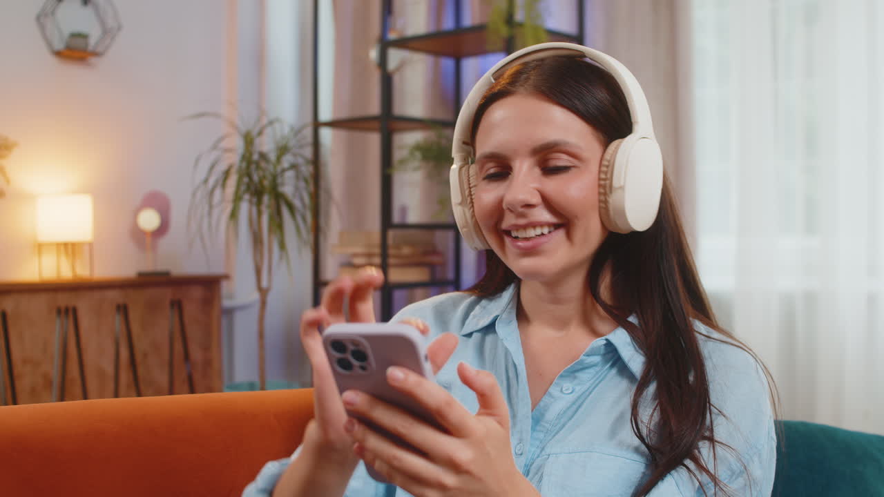 Giovane donna gioiosa che si diverte ad ascoltare musica con le cuffie wireless ballando sul divano della stanza di casa