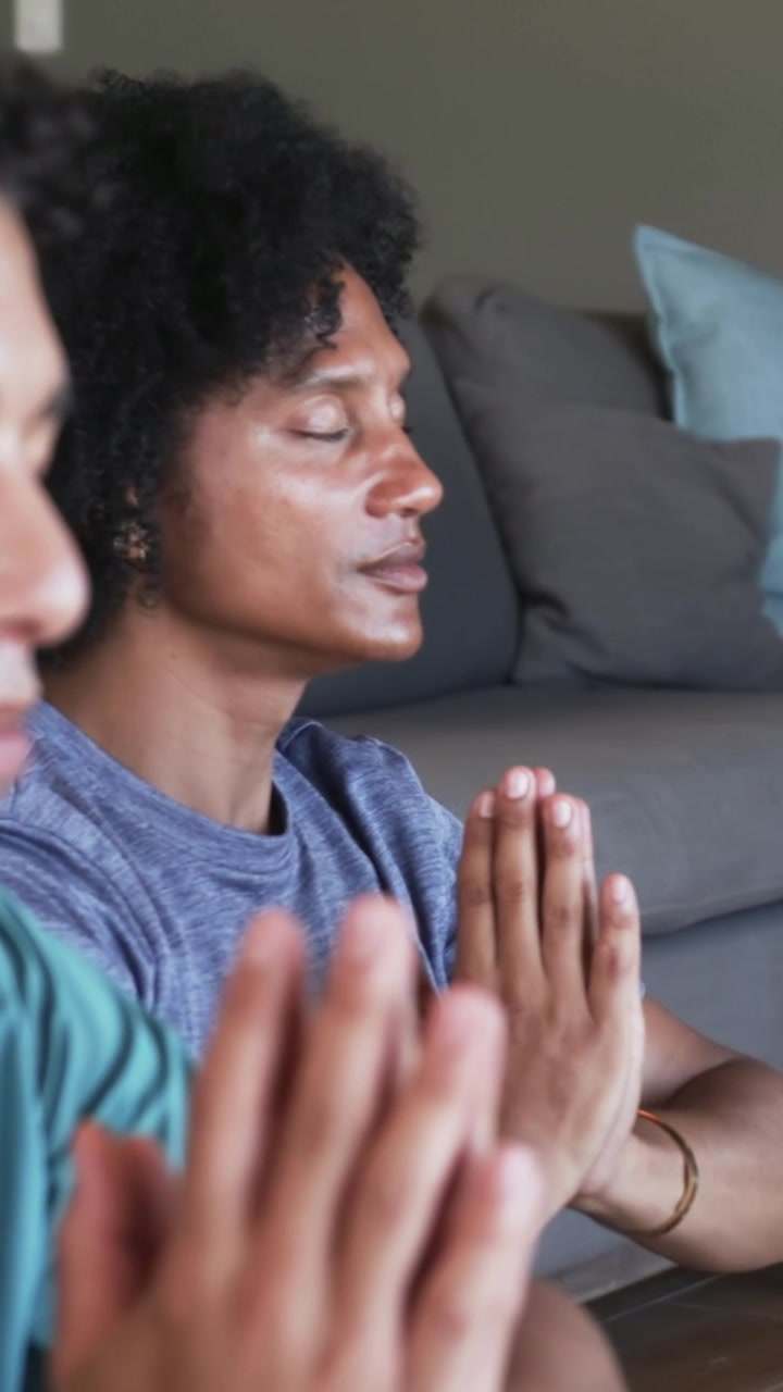 Vertical video: Closing eyes man in blue tee meditating hands pressed on floor at home near pillows