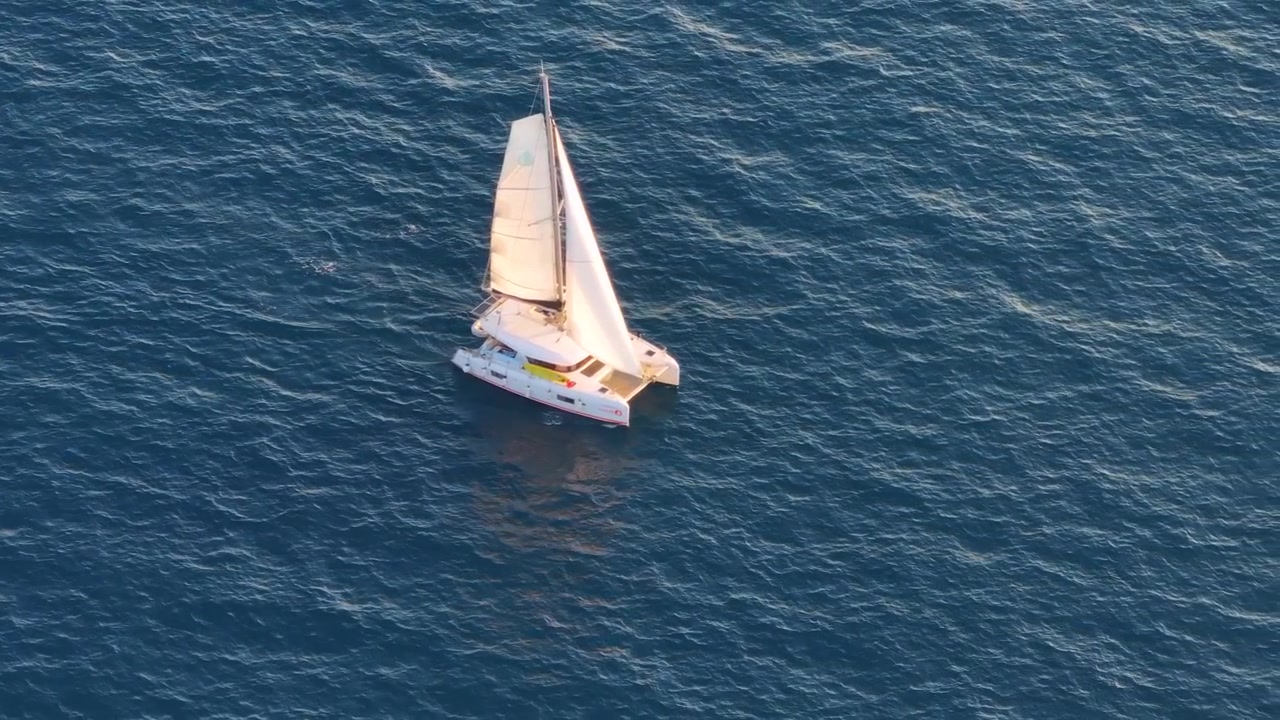 Una barca a vela naviga sull'acqua dell'oceano. La barca ha le vele alzate ed è in movimento. Il cielo è limpido e il sole illumina la scena. L'acqua appare calma